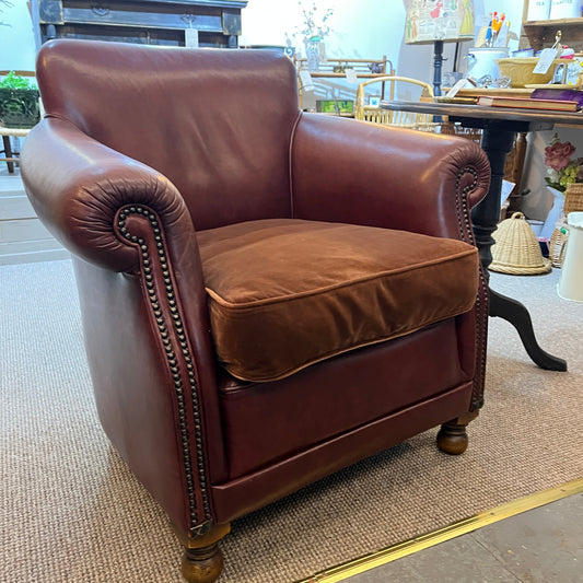 A classic Chesterfield-style leather armchair in reddish-brown, featuring rolled arms with nailhead trim, a matching cushion, and turned wooden legs, photographed in a furniture showroom.