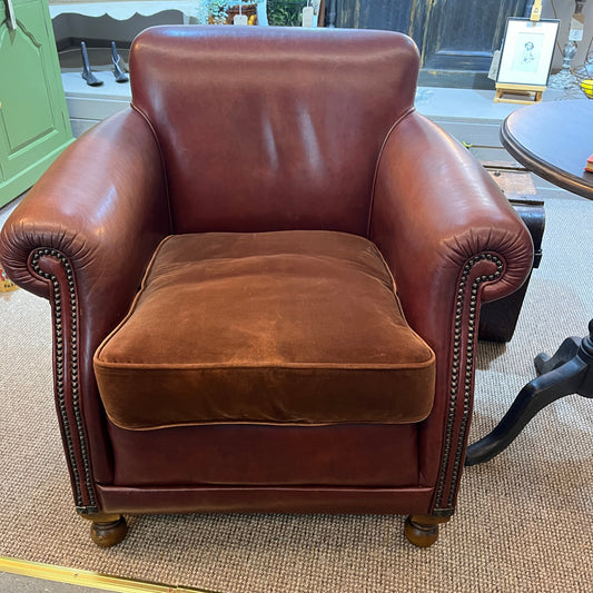 A classic brown leather armchair with rolled arms, nailhead trim, and a matching brown velvet seat cushion, photographed in a furniture showroom.