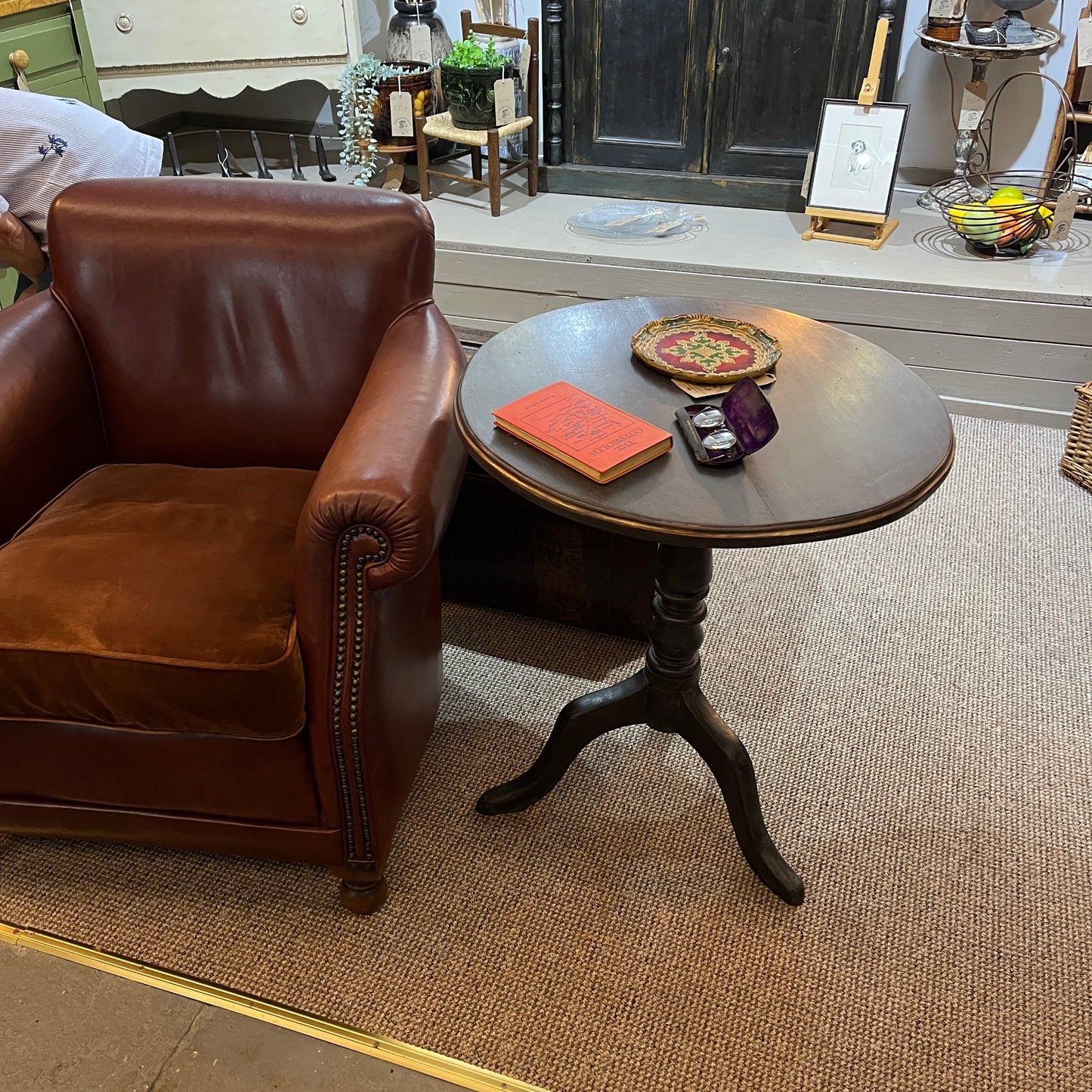 A brown leather armchair sits beside a small round wooden side table in a furniture showroom. The table has a dark pedestal base and holds a red book, a decorative plate, and a small purple pouch.