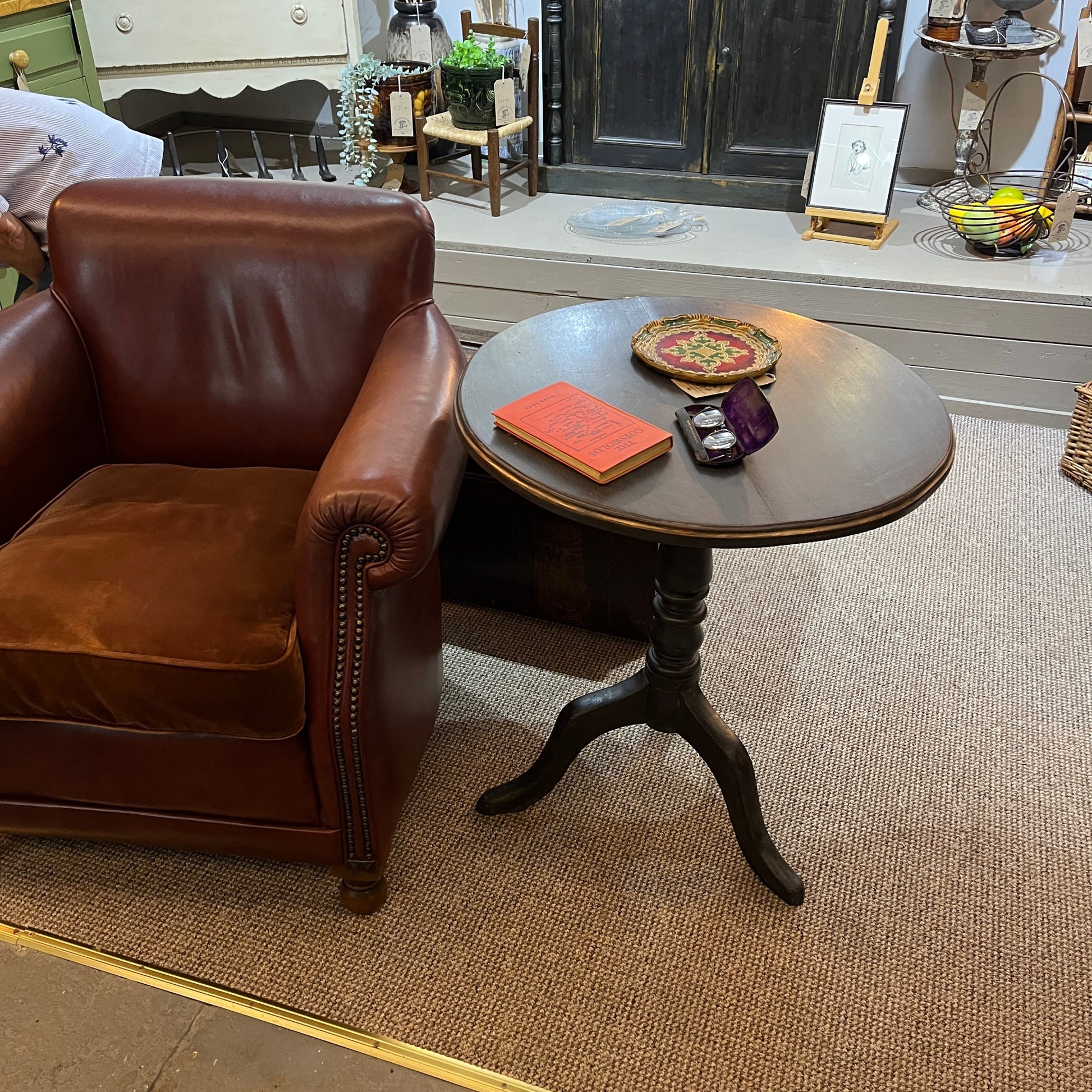 A brown leather armchair sits beside a small round wooden side table in a furniture showroom. The table has a dark pedestal base and holds a red book, a decorative plate, and a small purple pouch.