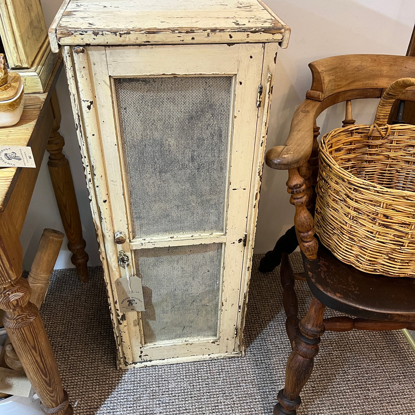 A distressed, vintage-style cream painted wooden cabinet with two mesh-screened doors, shown in a home decor setting next to a wooden chair with a wicker basket.