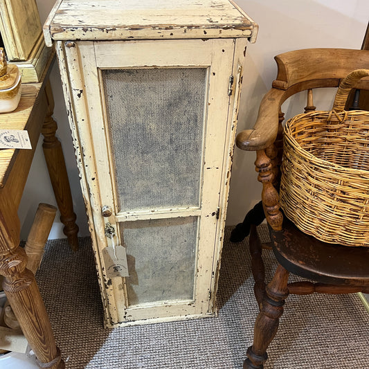 A distressed, vintage-style cream painted wooden cabinet with two mesh-screened doors, shown in a home decor setting next to a wooden chair with a wicker basket.