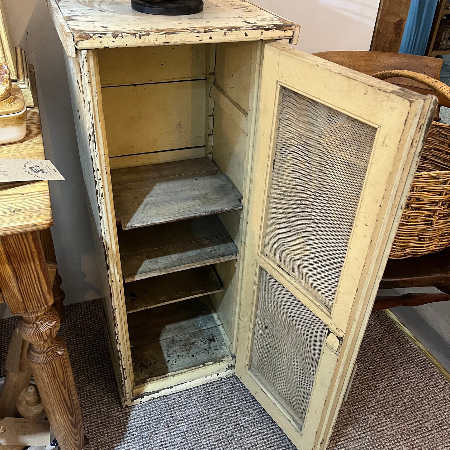 A distressed vintage wooden cabinet with a cream-colored frame and open door, showing three interior shelves, photographed in a home setting.