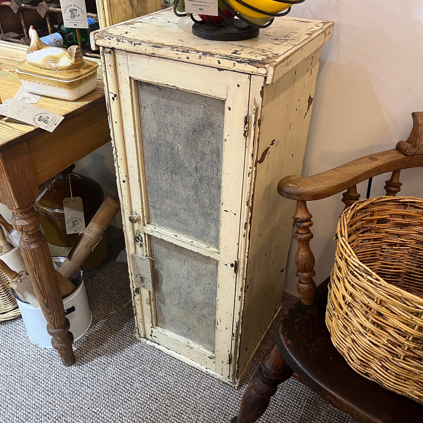 A distressed white wooden cabinet with two mesh screen windows, shown in a rustic shop or home setting next to a wooden chair and table.