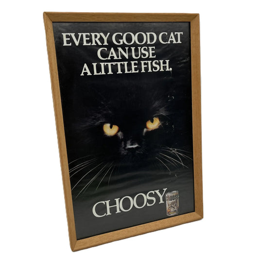 This is a vintage framed advertising poster for Choosy Cat Food. From the early 1980s, this one reads "every good cat can use a little fish". It has been framed in a wooden frame sometime in the past, which has helped to preserve it.