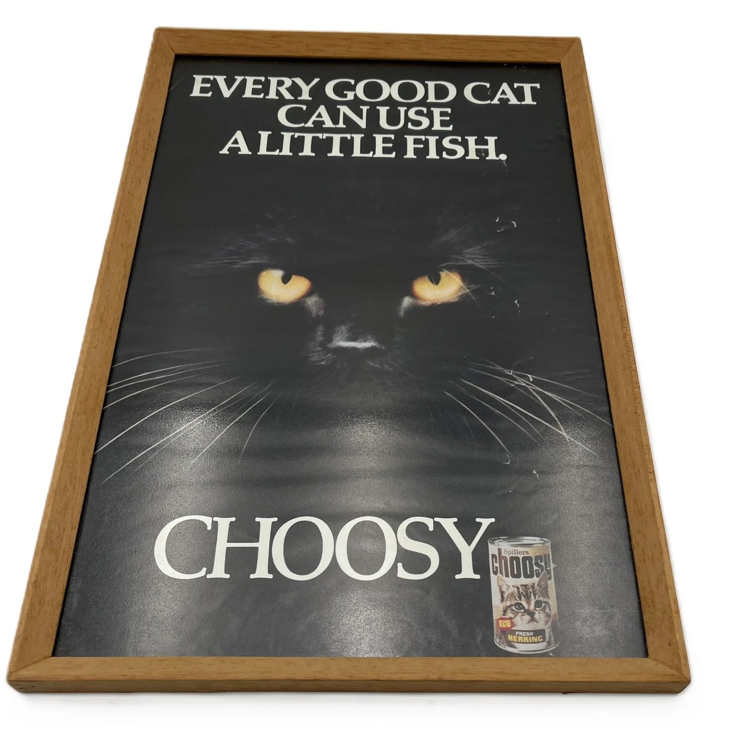 This is a vintage framed advertising poster for Choosy Cat Food. From the early 1980s, this one reads "every good cat can use a little fish". It has been framed in a wooden frame sometime in the past, which has helped to preserve it.