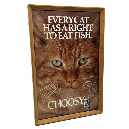 This is a vintage framed advertising poster for Choosy Cat Food. From the early 1980s, this one reads "every cat has the right to eat fish". It has been framed in a wooden frame sometime in the past, which has helped to preserve it.