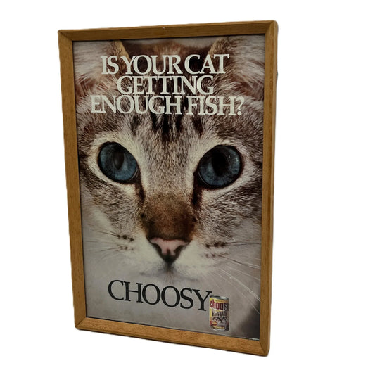 This is a vintage framed advertising poster for Choosy Cat Food. From the early 1980s, this one reads "is your cat getting enough fish". It has been framed in a wooden frame sometime in the past, which has helped to preserve it.