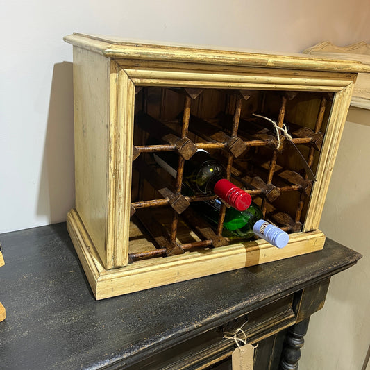 A rustic distressed wood wine rack cabinet with a cream-beige frame and brown wooden wine bottles inside, shown resting on a dark wooden table against a light wall.